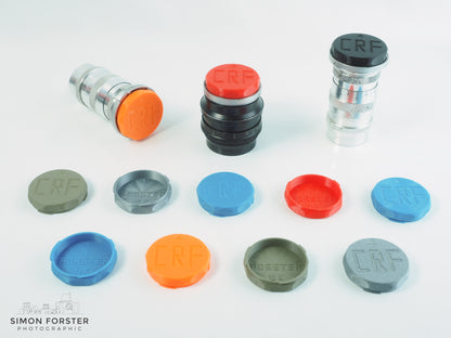 All colour variations of forster uk's CRF mount outer bayonet shallow rear lens caps are being displayed on a plain white background. The caps are being shown in black, orange, red, mid blue, silver and survival green, all which are available for purchase on simonforsterphotographic.co.uk.