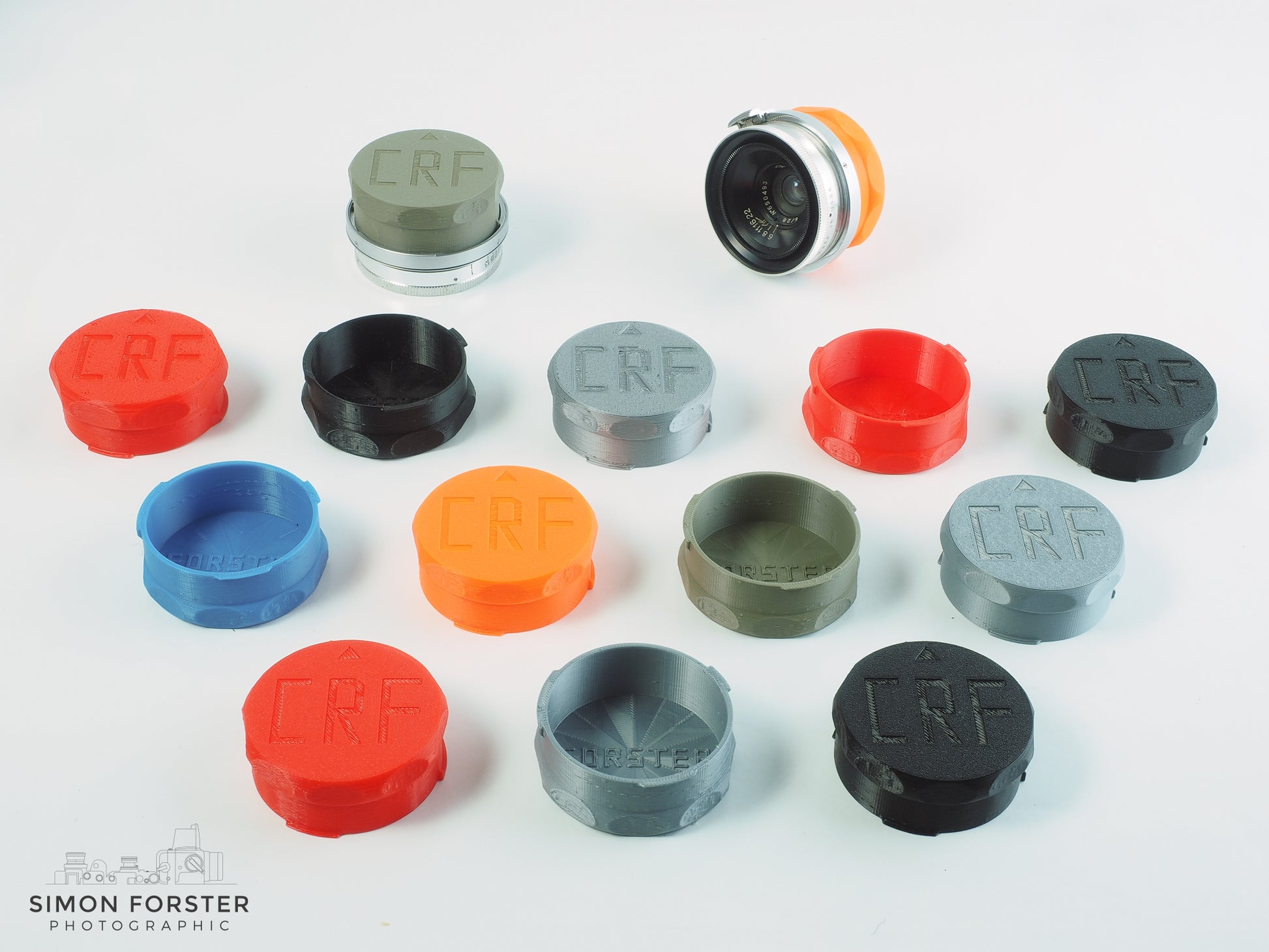 All colour variations of forster uk's CRF mount outer bayonet medium rear lens caps are being displayed on a plain white background. The caps are being shown in black, orange, red, mid blue, silver and survival green, all which are available for purchase on simonforsterphotographic.co.uk.