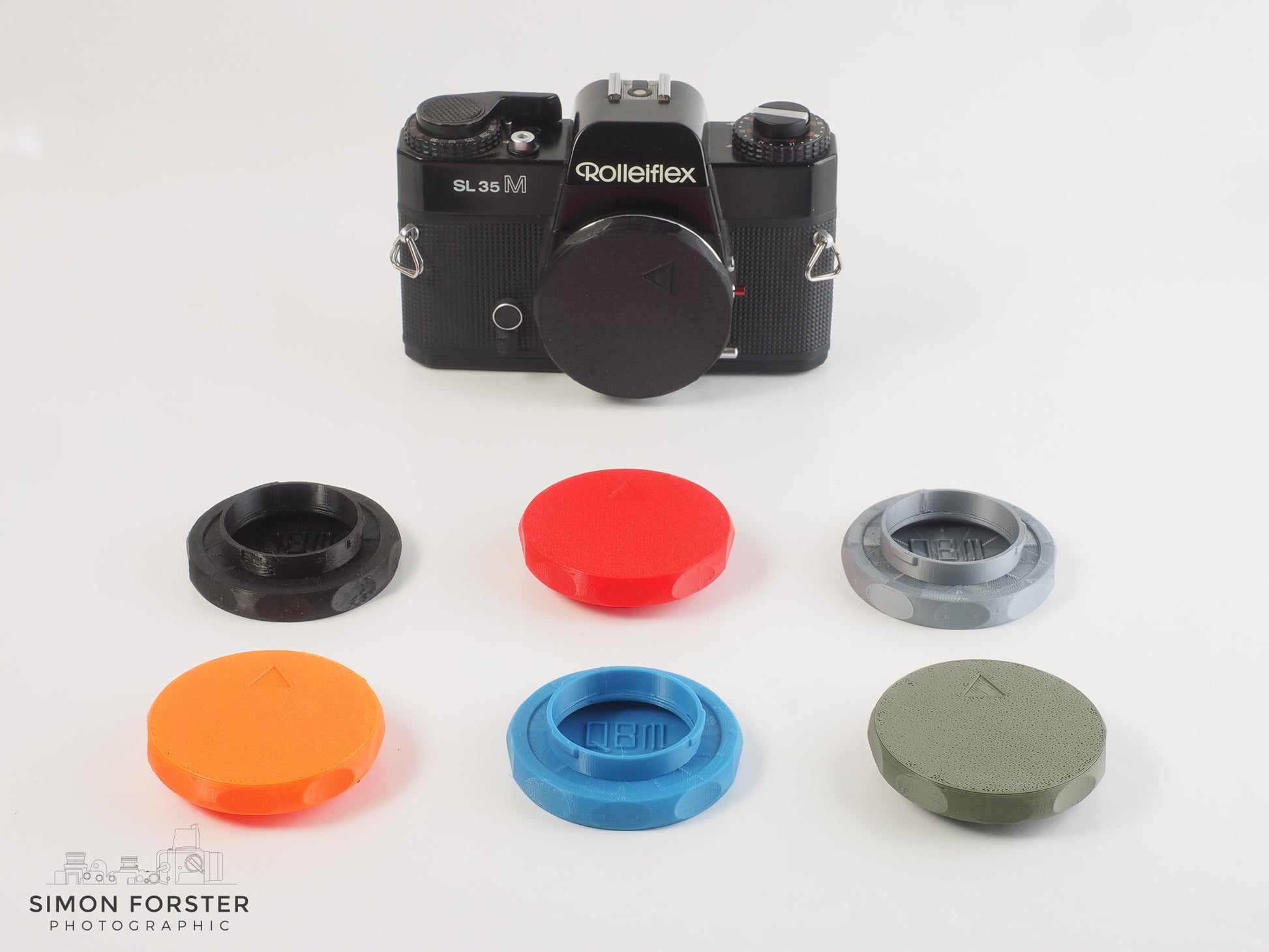 A collection of seven of the same Rolleiflex QBM body caps which all fit a Rolleiflex camera. The caps are in a range of colours, such as: Black, orange, red, light blue. silver and military green. Six of the caps are layed out together in two horizontal rows of three and the camera positioned behind the six caps with a black body cap being showcase on the Rolleiflex camera, the caps are displayed in a professional white background.