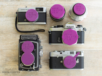 A collection of rear lens caps and camera body caps. The caps are bright purple and all say "#SHE HEARTS FILM" on the caps. There is a front cap which is fitted on a olympus trip 35 camera lens and camera, a front cap fitted onto a Rolleicord TLR camera, a body cap on a nikon camera, a rear lens cap fitting a lens with a m42 or m39 thread, and finally a rear cap fitted onto a exakta mount lens. The collection of caps are being displayed on a light timber organic background.