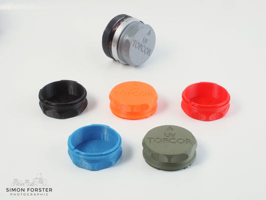 Six rear lens caps which have been designed to fit UV topcor lenses are being displayed on a plain white background including a UV topcor lens which has a silver cap mounted onto its rear element. The other caps are being showcase in front of the lens and silver cap. These caps are in black, orange, red, mid blue and survival green.